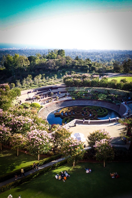 The image depicts a beautifully landscaped garden with a central circular water feature. The water feature is surrounded by a series of curved pathways and steps, leading to a central area with a maze-like design made of greenery. The garden is lush with various plants and trees, including flowering trees with pink blossoms. People are seen walking along the pathways, sitting on the grass, and enjoying the serene environment. In the background, a panoramic view of a cityscape is visible, with buildings and a clear blue sky. The overall atmosphere is peaceful and inviting, with a mix of natural and urban elements.</p>

<p>Provided by @altbot, generated privately and locally using Ovis2-8B