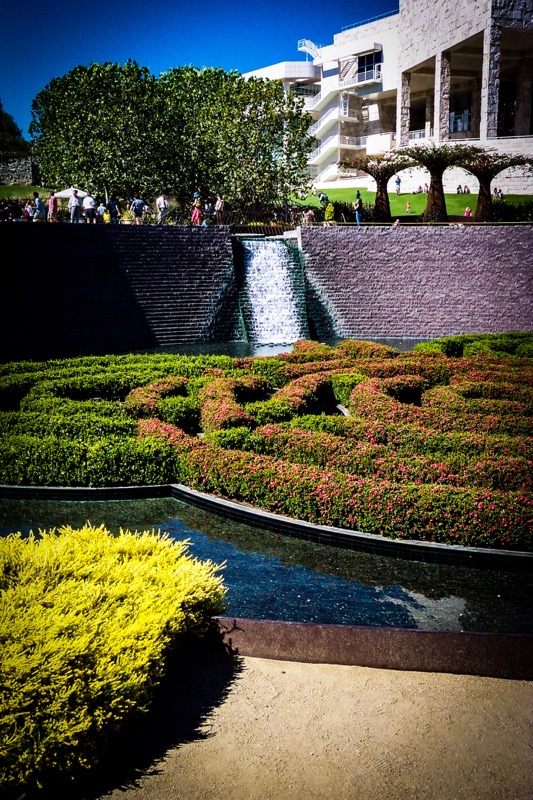 The image depicts a serene garden scene with a central waterfall cascading down a stone wall. In the foreground, there is a neatly trimmed hedge with vibrant green and pink flowers, and a reflective water feature that mirrors the surrounding greenery. The middle ground features a small waterfall with water flowing down a stepped stone wall, creating a tranquil ambiance. The background includes a modern building with white and gray stone façades, and several people are visible, some walking and others sitting on the grass. The sky is clear and blue, indicating a sunny day. The overall composition is well-balanced, with the waterfall as the focal point, surrounded by lush greenery and architectural elements.</p>

<p>Provided by @altbot, generated privately and locally using Ovis2-8B