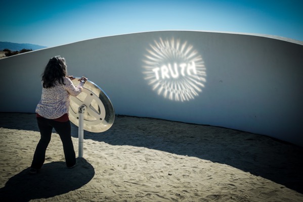 The image depicts an outdoor scene with a person interacting with a circular metallic device. The individual, facing away from the camera, is positioned on sandy ground, with a large white curved wall in the background. The person is holding a circular metallic object, which appears to be a mirror or a reflective surface, and is using it to project a bright light onto the wall. The light forms a circular pattern with the word TRUTH prominently displayed in the center. The sky is clear and blue, indicating a sunny day. The overall setting suggests an art installation or an interactive exhibit, with the focus on the projection of light and the word TRUTH.</p>

<p>Provided by @altbot, generated privately and locally using Ovis2-8B