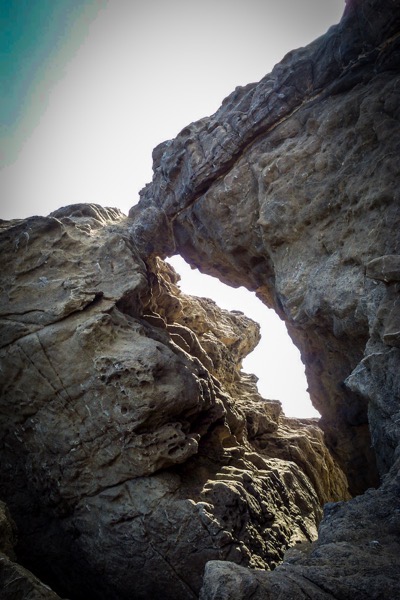 The image depicts a natural rock formation with a prominent archway. The rocks are rugged and weathered, displaying a range of earthy tones from light beige to dark gray. The archway is formed by the erosion of the rock, creating a natural opening that frames the sky. The texture of the rocks is rough, with visible layers and crevices, indicating geological processes over time. The lighting in the image suggests it was taken during the day, with sunlight illuminating the archway and casting shadows on the surrounding rock surfaces. The sky visible through the archway is a pale blue, contrasting with the darker tones of the rocks. The overall composition emphasizes the natural beauty and geological features of the landscape.</p>

<p>Provided by @altbot, generated privately and locally using Ovis2-8B