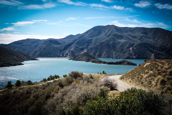 The image depicts a serene landscape featuring a large body of water surrounded by mountains. In the foreground, a winding road curves along the edge of the water, bordered by dry, shrubby vegetation. The mountains in the background are rugged and covered with sparse vegetation, with a prominent peak in the center. The sky is mostly clear with a few scattered clouds, and the water reflects the blue of the sky, creating a tranquil scene. The overall color palette includes shades of blue, green, and brown, contributing to the natural beauty of the setting.</p>

<p>Provided by @altbot, generated privately and locally using Ovis2-8B