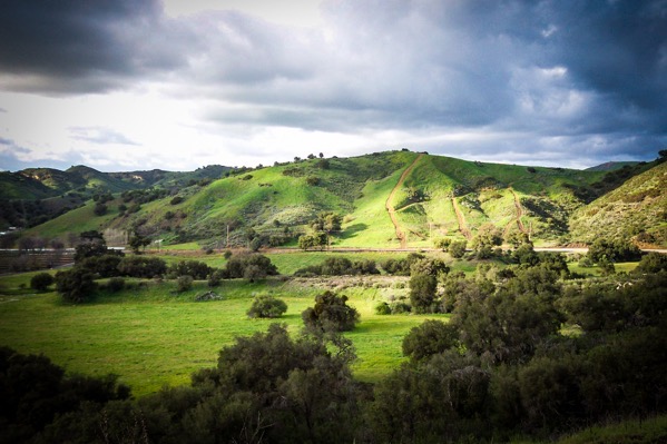  The image depicts a lush, green landscape with rolling hills under a dramatic sky. In the foreground, there is a grassy field dotted with small trees and shrubs, creating a textured appearance. The middle ground features a series of gently sloping hills, with paths or trails visible, suggesting human activity. The hills are covered in vibrant green grass, with some areas appearing darker due to shadows cast by the clouds. The sky is filled with dark, heavy clouds, contrasting with the bright sunlight that breaks through in some areas, highlighting the contours of the hills. The overall color palette is dominated by various shades of green, with the dark clouds adding depth and contrast.</p>

<p>Provided by @altbot, generated privately and locally using Ovis2-8B
