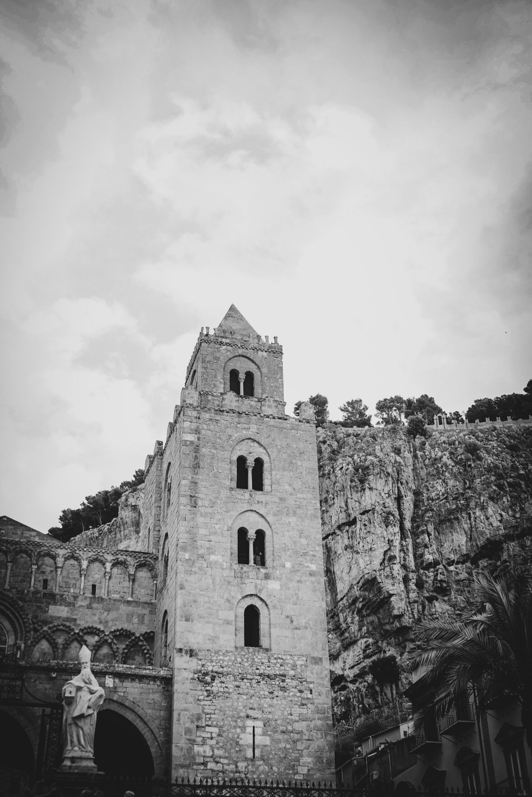  The image depicts a historic stone building, likely a church or cathedral, with a prominent bell tower. The structure is made of large, weathered stones, showcasing a mix of architectural styles, including Romanesque elements. The bell tower is tall and narrow, with arched windows and a pointed roof. The building is situated on a rocky cliff, with trees visible at the top, suggesting a natural, elevated setting. The sky is overcast, adding a dramatic effect to the scene. In the foreground, a statue of a religious figure stands near the entrance, adding to the historical ambiance. The photograph is in black and white, emphasizing the textures and contrasts of the stone and the surrounding landscape.</p>

<p>Provided by @altbot, generated privately and locally using Ovis2-8B