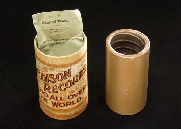 The image features a vintage Edison Records cylinder and its protective sleeve. The cylinder is cylindrical with a light brown color and a label that reads EDISON RECORDS WHO ALL OVER THE WORLD. The label is ornate, with decorative elements and text in a serif font. The top of the cylinder is open, revealing a green paper insert with the text No. 4705 Minstrel Record. Three Minutes with the Minstrels. The insert is partially pulled out, showing the text clearly. Next to the cylinder is a matching protective sleeve, also light brown, with a threaded top, indicating it was designed to fit over the cylinder. The background is black, providing a stark contrast to the objects.</p>

<p>Provided by @altbot, generated privately and locally using Ovis2-8B