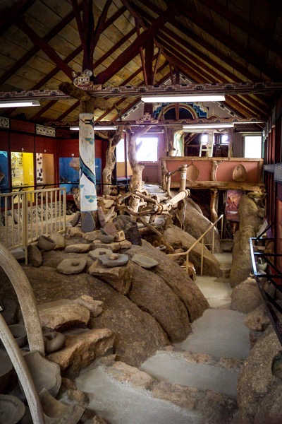 The image depicts an indoor archaeological exhibit with a rustic and historical ambiance. The space features a high, wooden ceiling with exposed beams and a decorative pattern along the edges. The walls are adorned with colorful, geometric patterns, enhancing the cultural theme. The floor is uneven, with large rocks and stones scattered throughout, creating a naturalistic setting. Various artifacts, including stone tools and pottery, are displayed on the ground, some partially buried, suggesting an excavation site. A large, carved wooden wheel is visible on the left side, adding to the historical context. The exhibit includes a raised platform with a railing, providing a vantage point to view the artifacts. Natural light filters in through windows, illuminating the space and highlighting the textures and colors of the exhibits. The overall layout and design suggest an immersive experience, allowing visitors to explore and learn about the historical context of the artifacts.</p></p>

<p><p>Provided by @altbot, generated privately and locally using Ovis2-8B