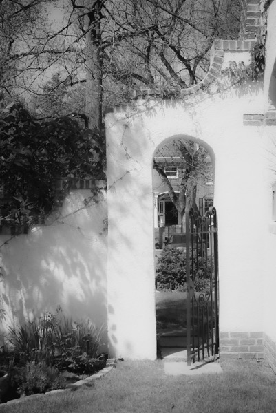 The image depicts a black and white scene featuring an arched gateway with a wrought iron gate. The gateway is part of a white wall with brick accents at the base. The gate is open, revealing a view of a garden and a house in the background. The garden includes various plants and shrubs, with a tree visible to the left of the arch. The house has a brick facade and a visible window. The scene is framed by bare tree branches, suggesting a winter or early spring setting. The sunlight casts shadows on the wall, adding depth to the image.</p>

<p>Provided by @altbot, generated privately and locally using Ovis2-8B
