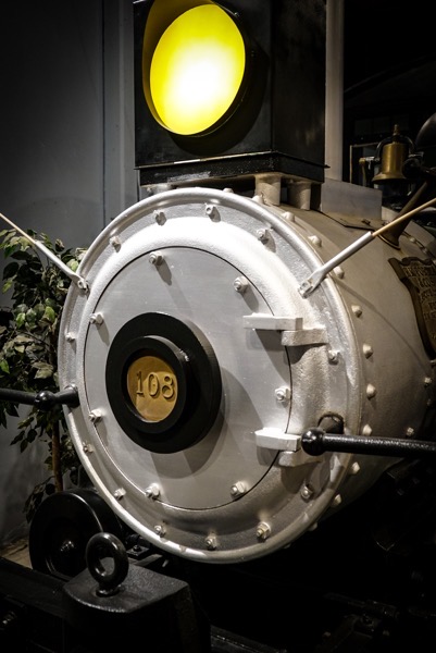The image features a close-up view of the front of a steam locomotive. The locomotives front is predominantly silver with a large, circular, metallic plate in the center, which has the number 108 prominently displayed in gold. The plate is secured with numerous bolts, and there are two black handles extending from the sides. Above the plate, a yellow circular light is visible, likely used for signaling. The background is dimly lit, with a plant visible to the left, adding a touch of greenery to the scene. The overall appearance suggests a well-maintained historical piece, possibly on display in a museum or exhibition.</p></p>

<p><p>Provided by @altbot, generated privately and locally using Ovis2-8B