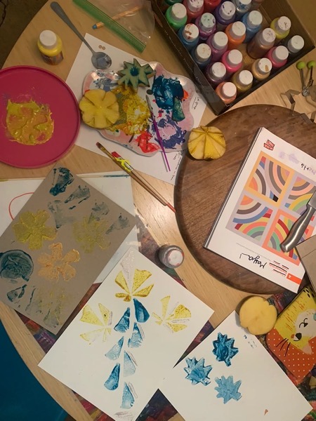 The image shows a cluttered table with various art supplies and materials. In the center, there is a wooden round tray with a knife and a book featuring colorful geometric patterns. Surrounding the tray are several sheets of paper with painted leaf-like shapes in yellow and blue. A palette with mixed paint and a spoon is visible, along with a pink plate with remnants of yellow paint and a halved pineapple used for stamping. To the right, a box of paint bottles in various colors is present. A paintbrush and a small bottle of yellow paint are also on the table. The overall scene suggests an ongoing art project involving stamping techniques.</p>

<p>Provided by @altbot, generated privately and locally using Ovis2-8B