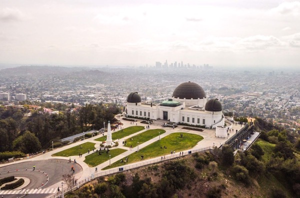 The image depicts the Griffith Observatory, a prominent landmark in Los Angeles, California. The observatory is characterized by its distinctive white architecture and two large domes, one of which is a prominent feature. The building is situated on a hill, offering a panoramic view of the city below. The foreground shows a well-maintained lawn with pathways and a few visitors scattered across the area. A circular road is visible, leading up to the observatory. In the background, the sprawling cityscape of Los Angeles is visible, with the downtown skyline faintly discernible in the distance. The sky is overcast, with a hazy atmosphere, suggesting a typical day in the city. The image captures the observatory's significance as a cultural and scientific landmark, with its location providing a vantage point over the urban landscape.</p>

<p>Provided by @altbot, generated privately and locally using Ovis2-8B