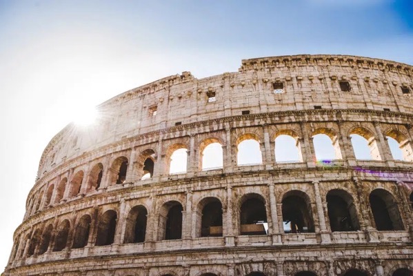 The image depicts the Colosseum, an iconic ancient amphitheater in Rome, Italy. The structure is shown from a low angle, emphasizing its grandeur and architectural details. The sun is positioned to the left, casting a warm glow and creating a lens flare effect. The Colosseum's weathered stone facade is visible, with multiple levels of arches and openings. The sky is clear and blue, enhancing the contrast with the structure. The image captures the Colosseum's historical significance and architectural beauty.</p>

<p>Provided by @altbot, generated privately and locally using Ovis2-8B