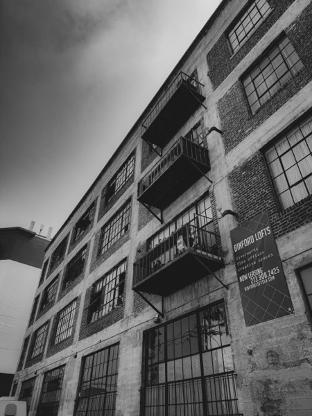 The image depicts a multi-story brick building with a monochromatic, black-and-white aesthetic. The building features large, rectangular windows with metal grills, and several fire escapes with metal railings extend from the second to the fourth floors. The facade is a mix of exposed brick and concrete, with some areas showing signs of wear and age. A sign on the right side of the building reads "BINFORD LOFTS" and provides information about live/work spaces, flex offices, and creative spaces, along with a phone number and website. The sky is overcast, adding to the industrial feel of the scene. The building is situated in an urban environment, with another structure visible in the background.</p>

<p>Provided by @altbot, generated privately and locally using Ovis2-8B