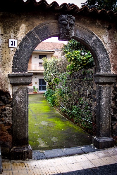 The image features a stone archway with a carved face at the top, leading to a narrow pathway. The archway is part of an old, weathered wall with a number 31 displayed on it. The pathway is covered with green moss, indicating dampness, and is bordered by a stone wall on the right, which is partially covered with greenery. In the background, a two-story building with a red-tiled roof and a balcony is visible. The building has a blue door and windows, and there are potted plants near the entrance. The scene is set in a residential area, with a mix of natural and man-made elements.</p>

<p>Provided by @altbot, generated privately and locally using Ovis2-8B