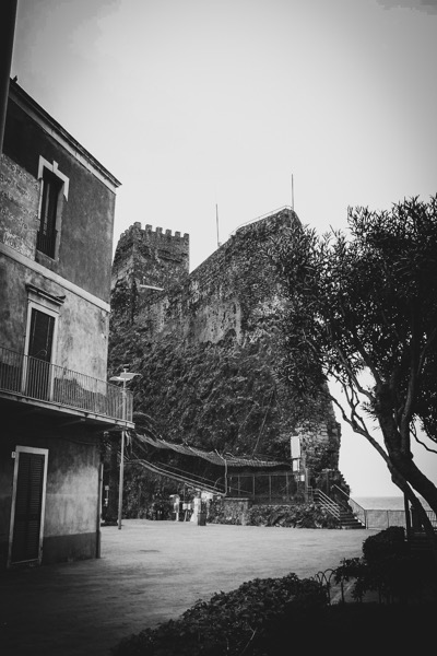 The image depicts a historical scene featuring a stone castle with a prominent tower, set against a backdrop of a clear sky. The castle is built on a rocky outcrop, with visible crenellations on the tower, suggesting its defensive purpose. Adjacent to the castle is a two-story building with a balcony and a door, displaying signs of age and weathering. The foreground includes a paved area with a tree on the right, adding a touch of greenery to the scene. The photograph is in black and white, enhancing the historical ambiance. The overall composition highlights the contrast between the ancient stone structures and the natural elements, such as the tree and the sky.