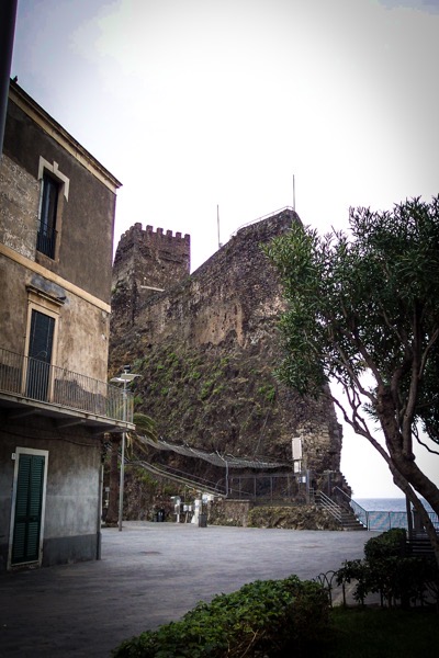 The image depicts a historical scene featuring a large stone fortress with a prominent tower, set against a backdrop of a cloudy sky. The fortress is built into a rocky cliff, with a staircase leading up to it, suggesting it was once a defensive structure. In the foreground, there is a building with a balcony and green shutters, indicating a residential or commercial use. The building's facade is weathered, with visible signs of age and wear. A tree with green leaves is visible on the right side of the image, adding a touch of nature to the scene. The ground is paved, and there are some green bushes and a metal fence near the base of the fortress. The ocean is visible in the background, suggesting the location is coastal. The overall atmosphere is serene and historical, with a focus on the architectural elements and natural surroundings.</p></p>

<p><p>Provided by @altbot, generated privately and locally using Ovis2-8B