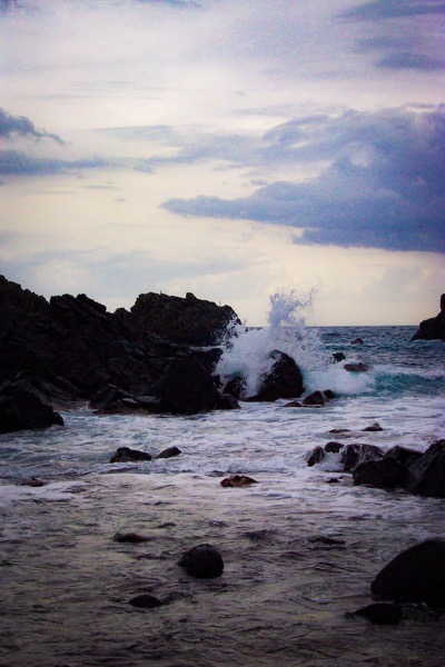The image depicts a coastal scene with waves crashing against dark, jagged rocks. The foreground features a rocky shoreline with various-sized rocks scattered across the water's surface. The waves are white and frothy, creating a dynamic splash as they hit the rocks, with the water appearing to be a deep blue-green color. The background shows a vast expanse of the ocean meeting a cloudy sky, with the clouds appearing gray and overcast, suggesting a potentially stormy atmosphere. The overall lighting is dim, with the scene being illuminated by a soft, diffused light, possibly indicating early morning or late afternoon. The image captures the raw power and beauty of the natural coastal environment.</p></p>

<p><p>Provided by @altbot, generated privately and locally using Ovis2-8B