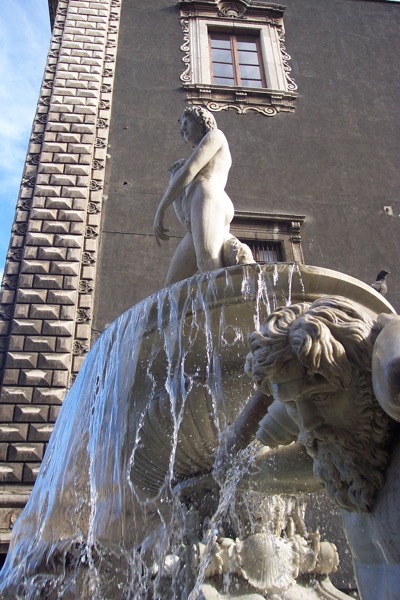 The image depicts a classical fountain with a statue of a nude figure standing atop a large, round basin. The statue, likely representing a mythological or historical figure, is positioned with its back to the viewer, gazing downward. Water cascades over the edge of the basin, creating a dynamic flow that adds movement to the scene. The fountain is intricately designed, with a sculpted figure at the base, possibly a sea creature, from which water emerges. The background features a building with ornate architectural details, including a window with decorative trim. The photograph is in black and white, emphasizing the textures and contrasts between the stone of the statue and the flowing water. A bird is perched on the edge of the fountain, adding a touch of life to the scene.</p>

<p>Provided by @altbot, generated privately and locally using Ovis2-8B