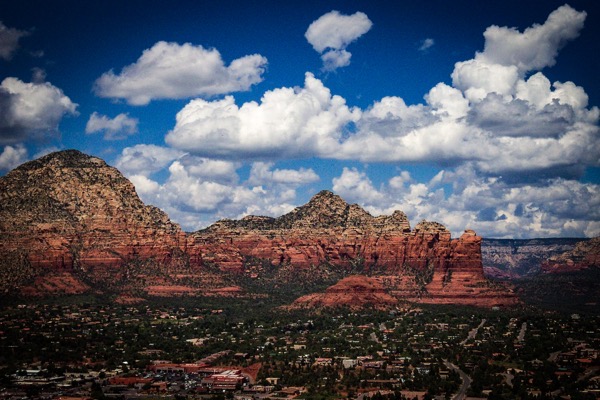 The image depicts a scenic view of a town nestled in a valley with a prominent red rock formation in the background. The sky is a vibrant blue with scattered white clouds, creating a striking contrast with the earthy tones of the landscape. The red rock formation, characterized by its layered sedimentary rock, rises majestically above the town, showcasing a rugged and textured surface. The town itself is visible in the foreground, with a mix of residential and commercial buildings, roads, and greenery, indicating a blend of natural and human-made elements. The overall composition highlights the grandeur of the natural landscape juxtaposed with the presence of human habitation.</p></p></p>

<p><p><p>Provided by @altbot, generated privately and locally using Ovis2-8B