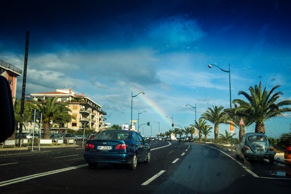 The image depicts a street scene with a clear blue sky and a rainbow visible in the background. In the foreground, a blue car is driving on a multi-lane road, with a license plate reading "BP 455EM." The road is lined with palm trees on the right side, and there are buildings on the left, including a multi-story structure with balconies. Streetlights are visible along the road, and traffic signs are present, indicating a curve ahead. The scene is well-lit, suggesting it is daytime. The rainbow adds a vibrant touch to the otherwise urban setting.</p>

<p>Provided by @altbot, generated privately and locally using Ovis2-8B