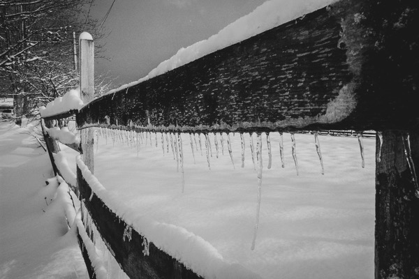 The image depicts a winter scene with a wooden fence covered in snow and icicles. The fence, made of dark wood, is partially obscured by a thick layer of snow on top. Icicles hang from the bottom edge of the fence, varying in length and thickness, indicating freezing temperatures. The snow on the ground is undisturbed, suggesting a calm and quiet environment. In the background, there are bare trees and a power line, adding to the wintry atmosphere. The photograph is in black and white, enhancing the contrast between the dark fence and the bright snow.</p></p></p></p></p>

<p><p><p><p><p>Provided by @altbot, generated privately and locally using Ovis2-8B