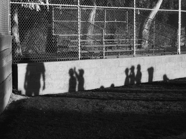 The image is a black and white photograph capturing the shadows of people cast on a wall. In the foreground, the shadows of at least five individuals are visible, with one appearing larger, suggesting an adult, and the others smaller, indicating children. The shadows are elongated, indicating the light source is low, likely the sun setting or rising. The background features a chain-link fence with metal bleachers behind it, suggesting a sports field or park setting. The ground is grassy, and the wall is made of concrete blocks. The shadows are crisp and well-defined, contrasting sharply with the lighter background.</p></p>

<p><p>Provided by @altbot, generated privately and locally using Ovis2-8B