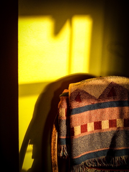 The image features a colorful woven blanket draped over a chair, with sunlight casting a warm yellow glow on the wall behind it. The blanket has a pattern of geometric shapes, including triangles and squares, in shades of red, blue, and beige. The sunlight creates a distinct shadow of the chair and blanket on the wall, enhancing the texture and colors of the blanket. The scene is set in a dimly lit room, with the sunlight coming from the left side, creating a contrast between the illuminated and shadowed areas. The overall atmosphere is cozy and inviting, with the blanket's texture and the interplay of light and shadow adding depth to the image.</p></p>

<p><p>Provided by @altbot, generated privately and locally using Ovis2-8B