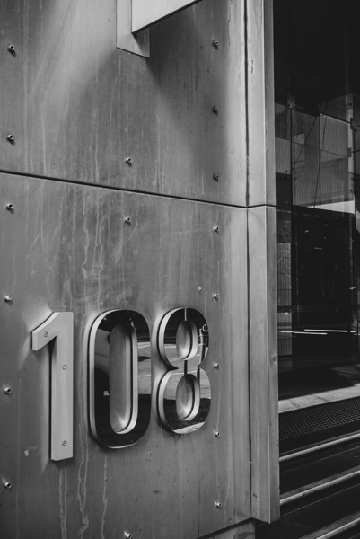 The image is a close-up, monochromatic shot of a building's exterior featuring large, metallic numerals. The numbers "108" are prominently displayed, appearing raised and three-dimensional against a textured, dark gray background. The background consists of vertically aligned, rectangular metal panels secured with visible screws or bolts. To the right of the numbers, a partially visible doorway or entryway suggests depth and leads into a darker space with what looks like a metal grate. The overall composition is industrial and minimalist, with a focus on texture and form.</p>

<p>Provided by @altbot, generated privately and locally using Gemma3:27b