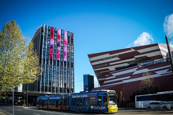 A daytime outdoor shot depicts a street scene with modern architecture and public transportation. On the left is a tall, rectangular building with a facade of many narrow, vertical windows, displaying vibrant, colorful patterns on some of the windows. A blue tram with the text "Know how to grow" is positioned in front of the building, and the logo "UniSuper" is prominently displayed on its side. To the right of the tram is a multi-faceted, reddish-brown building labeled "ADELAIDE CONVENTION CENTRE". The sky is clear blue with a few scattered white clouds, and a bus is partially visible on the right side of the frame.</p>

<p>Provided by @altbot, generated privately and locally using Gemma3:27b