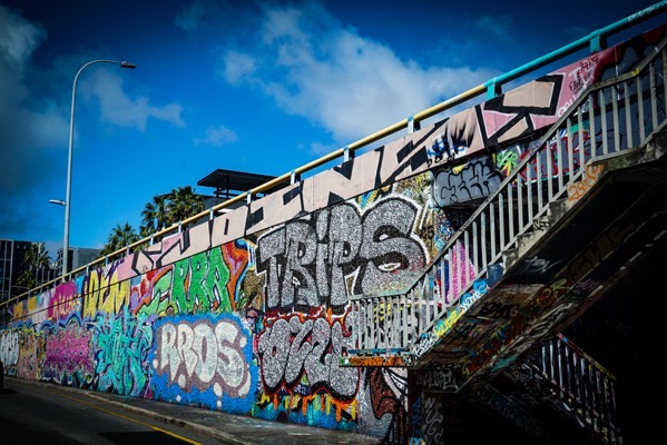 A long, angled wall covered in vibrant graffiti art is shown from a low perspective, extending upwards and to the right. The wall is a patchwork of colorful spray paint in various styles and patterns, creating a dense, chaotic visual texture. A metal railing with vertical bars runs along the top of the wall, following its incline. The foreground shows a dark asphalt road, contrasting with the bright colors of the graffiti. Parts of the wall are obscured by shadows, and the sky above is a clear blue with some scattered clouds. The word PROS is clearly visible in white and gray paint on the wall.</p>

<p>Provided by @altbot, generated privately and locally using Gemma3:27b
