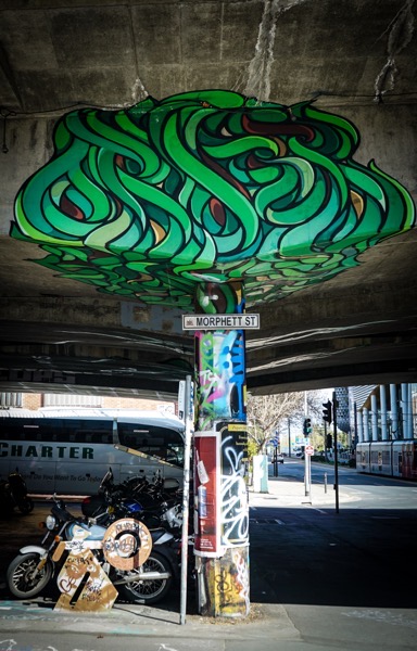 A photograph depicts an urban underpass with a complex mural painted on the ceiling. The mural consists of interwoven green and blue shapes, resembling stylized leaves or plant forms. Below the mural, a street sign reads MORPHETT ST is affixed to a cylindrical concrete support beam. To the left, a motorcycle is parked next to several cardboard pieces covered in spray paint. The underpass extends into the distance, revealing a street scene with traffic signals and buildings, indicating an active city environment.</p>

<p>Provided by @altbot, generated privately and locally using Gemma3:27b