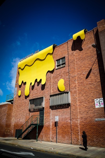 A large brick building with a striking yellow paint design resembling dripping liquid covers a significant portion of the facade. The building has several windows of varying sizes, and a dark metal staircase leading up to a doorway on the left side. A small, rectangular sign reading “ONE WAY” is visible beneath one of the windows. To the right of the building, a small square poster with pink and white imagery is affixed to the brick. Text on the top of the building reads LION ARTS CENTER.</p>

<p>Provided by @altbot, generated privately and locally using Gemma3:27b