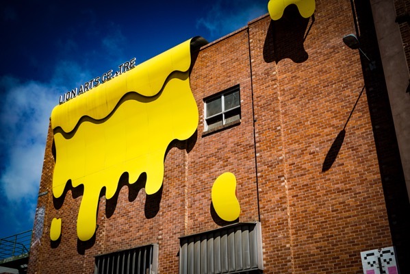 A wide shot of a multi-story brick building with a large, dripping yellow paint mural on its side. The building is red-brown in color and features several rectangular windows, some of which are covered by metal grates. The mural depicts a large, amorphous shape of dripping yellow paint, resembling melted cheese or flowing liquid, which covers a significant portion of the building's facade. The paint appears to be spilling over the edges of the building. Visible text on the mural reads LION ARTS CENTER. A portion of a dark blue sky is visible in the background.</p>

<p>Provided by @altbot, generated privately and locally using Gemma3:27b