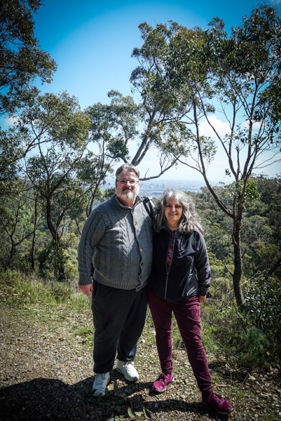 Two people are standing outdoors in a natural setting, likely a hillside or wooded area. The person on the left is wearing a gray cable-knit sweater, dark pants, and white sneakers. The person on the right is wearing a black jacket with red accents, maroon pants, and purple shoes. The hillside is covered in green shrubbery, and trees surround them on either side. In the background, a distant landscape of hills and a bright blue sky are visible.</p></p>

<p><p>Provided by @altbot, generated privately and locally using Gemma3:27b
