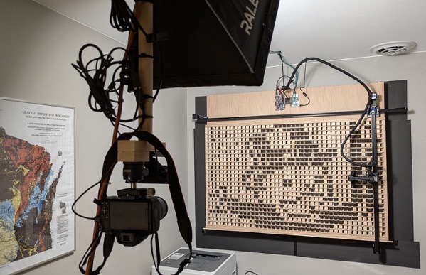 This image depicts a recording studio setup, likely for voice-over or audio production. The main focus is on a complex microphone array mounted on a wall, consisting of a grid of numerous small microphones attached to a wooden panel with dark gray framing. To the left of the microphone array is a large camera mounted on a tripod, with numerous cables running from it and around the studio.</p></p>

<p><p>Provided by @altbot, generated privately and locally using Gemma3:27b